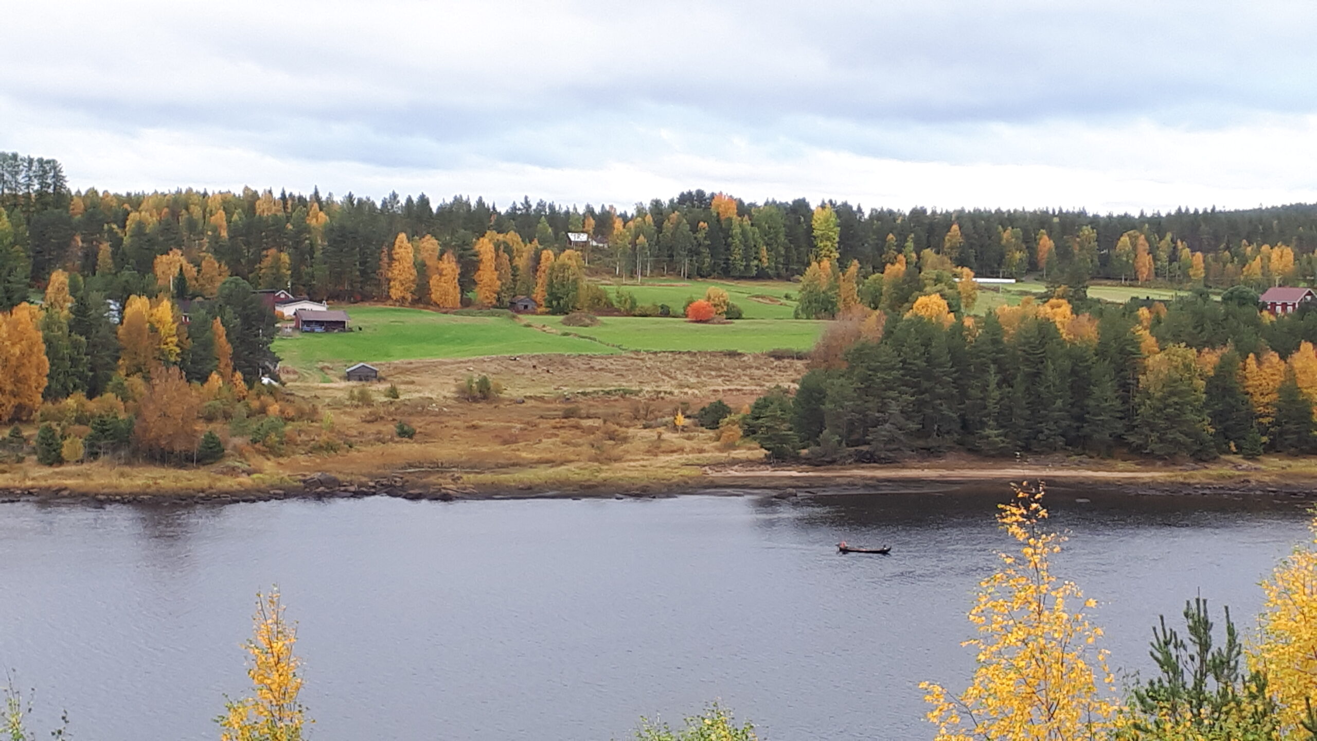 Maalaismaisemassa joella kalastaja veneessä ja taustalla rinteessä peltoja ja maalaistaloja